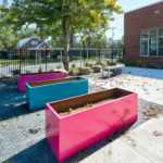 Three large, empty, colorful rectangular planters sit on gravel near a brick building, with a black metal fence and trees in the background.