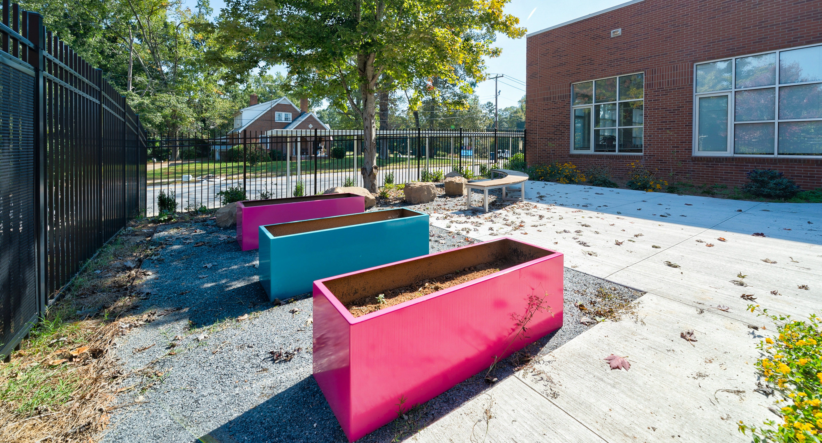 maglin_mpl-1500-planters-2 Three large, empty, colorful rectangular planters sit on gravel near a brick building, with a black metal fence and trees in the background.