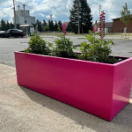 A bright pink rectangular planter with green plants and pink flowers is placed on a sidewalk beside a parking lot and buildings under a partly cloudy sky.