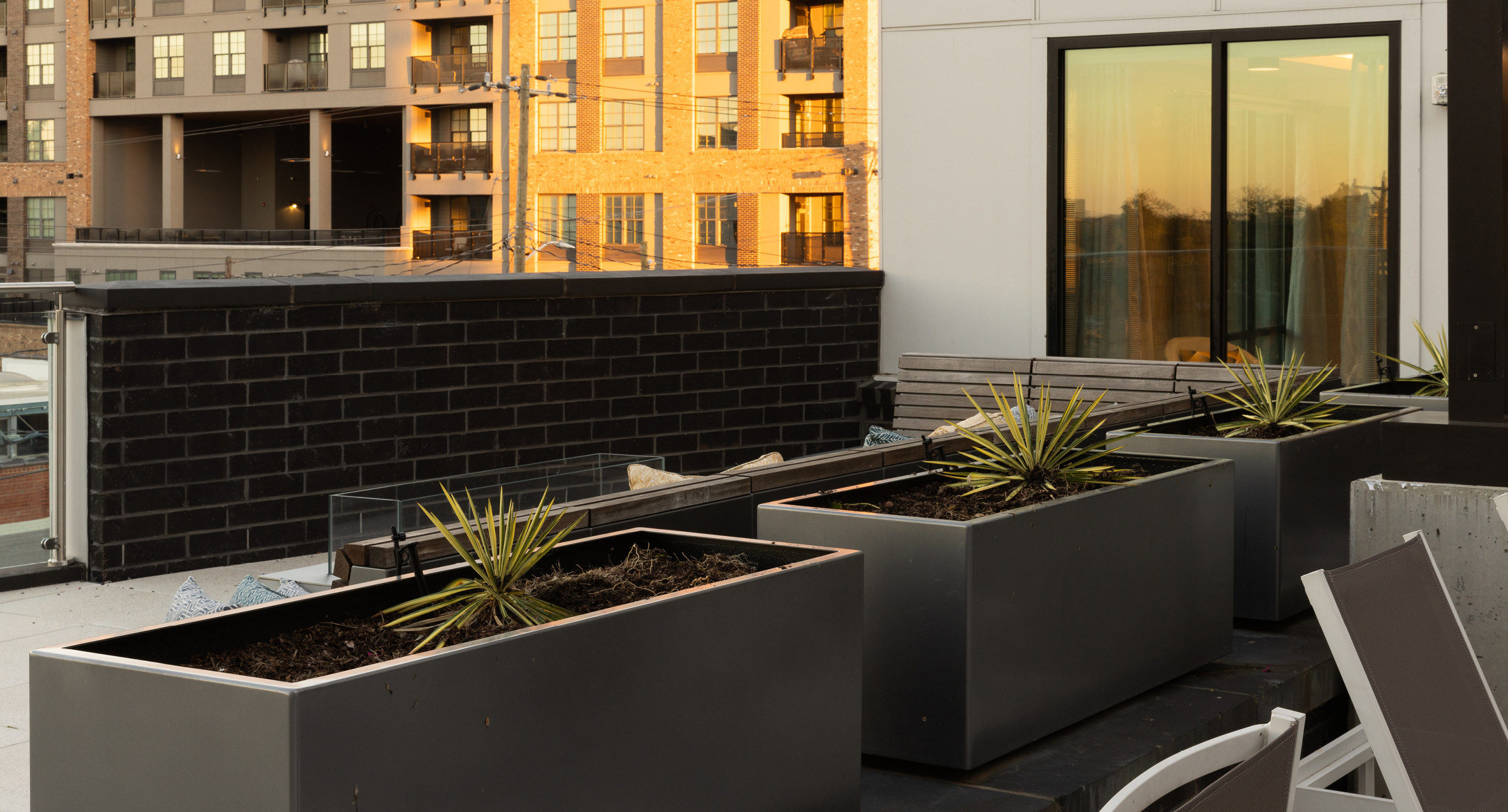 maglin_mpl-1500-planters-4 Three rectangular black planters with small spiky plants are arranged on a modern rooftop patio, with buildings and sliding glass doors in the background.