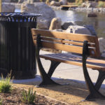Un banc public en bois est assis à côté d'une poubelle noire près d'un étang, avec des rochers, un trottoir et des plantes en arrière-plan.