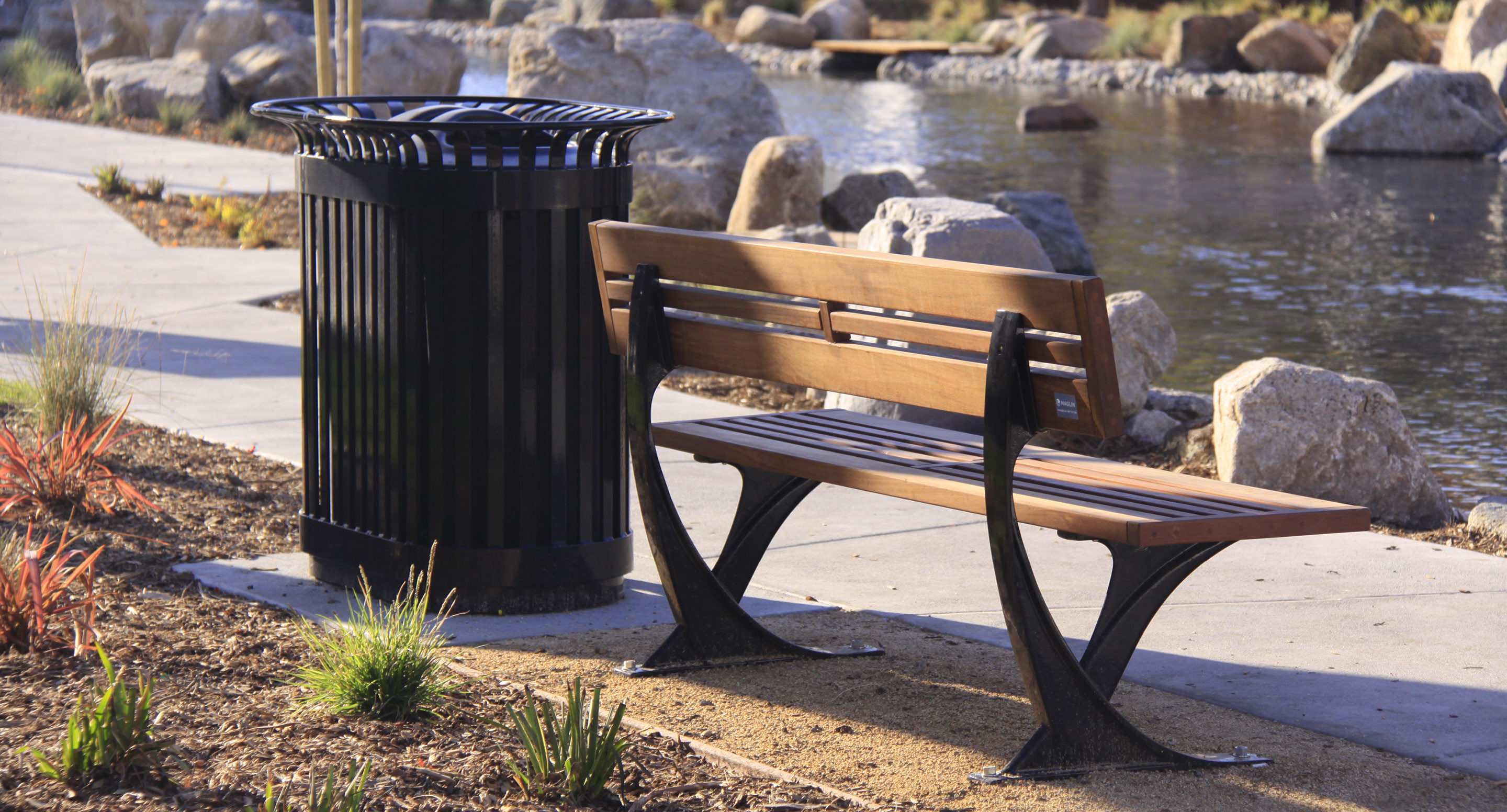 maglin_mrr-0200-recycle-receptacles-0611 Un banc public en bois est assis à côté d'une poubelle noire près d'un étang, avec des rochers, un trottoir et des plantes en arrière-plan.