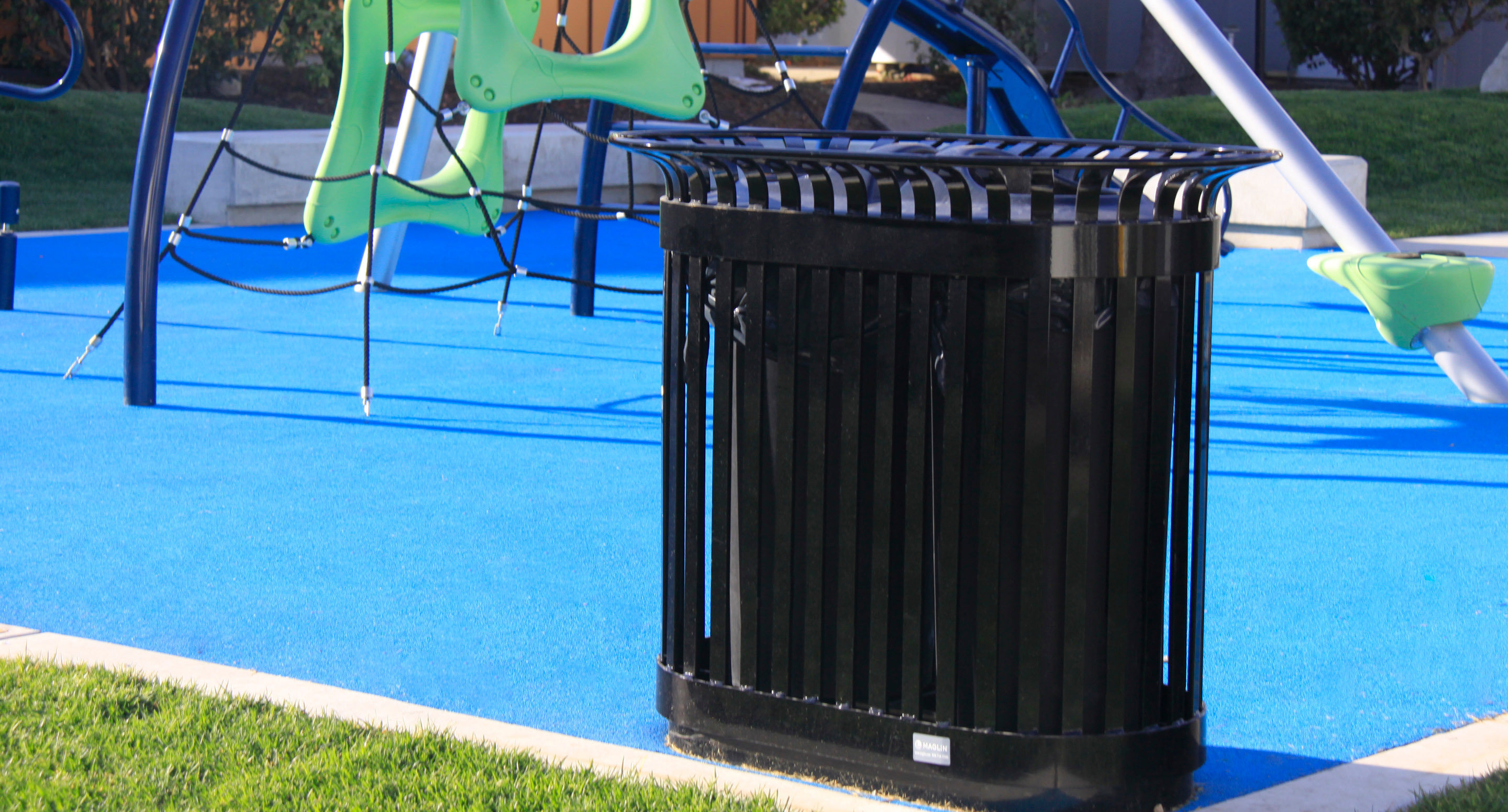 maglin_mrr-0200-recycle-receptacles-1 Une poubelle en métal noir se trouve sur le bord d'un terrain de jeu en caoutchouc bleu avec des équipements d'escalade en arrière-plan.