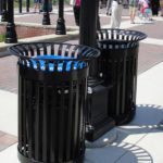 Deux poubelles métalliques noires sont placées côte à côte sur un trottoir pavé, près d'un lampadaire, dans un espace public en plein air, avec des personnes en arrière-plan.