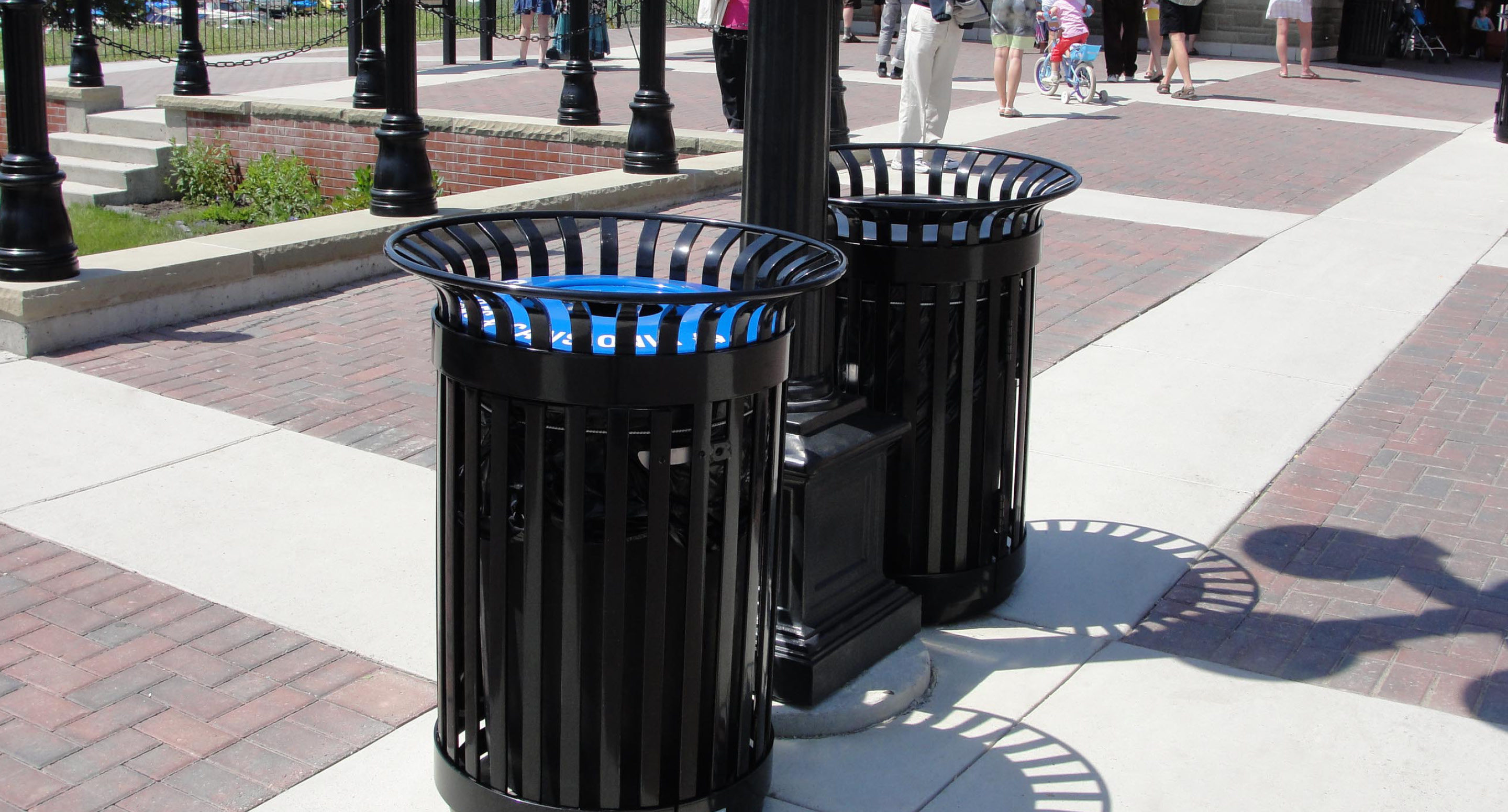 Deux poubelles métalliques noires sont placées côte à côte sur un trottoir pavé, près d'un lampadaire, dans un espace public en plein air, avec des personnes en arrière-plan.