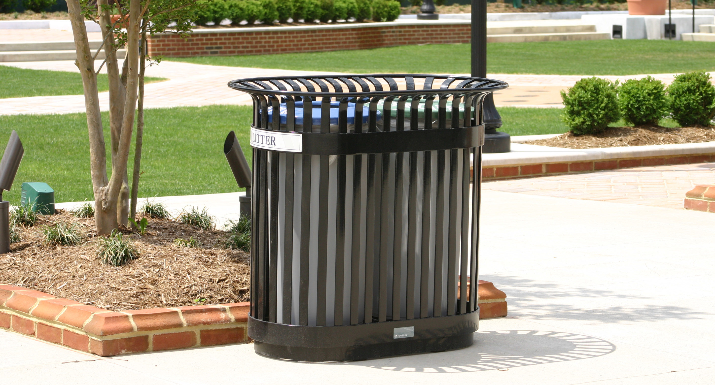 maglin_mrr-0200-recycle-receptacles-4 Une poubelle en métal noir est placée sur un trottoir près d'une zone paysagée avec de l'herbe, des buissons et des bordures en briques.