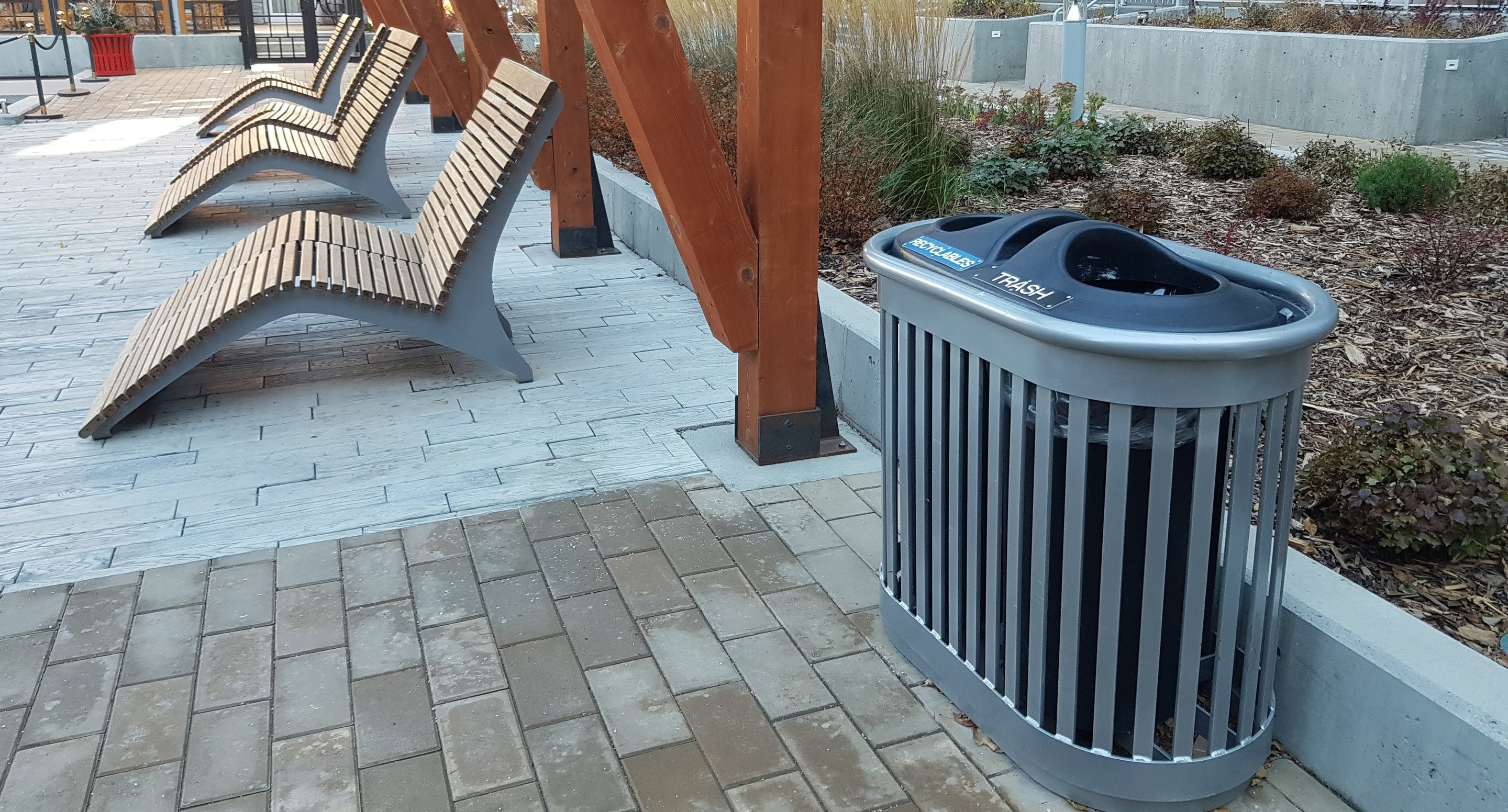 Plusieurs chaises longues en bois et une poubelle étiquetée sur un patio extérieur pavé avec un aménagement paysager et des poteaux en bois.