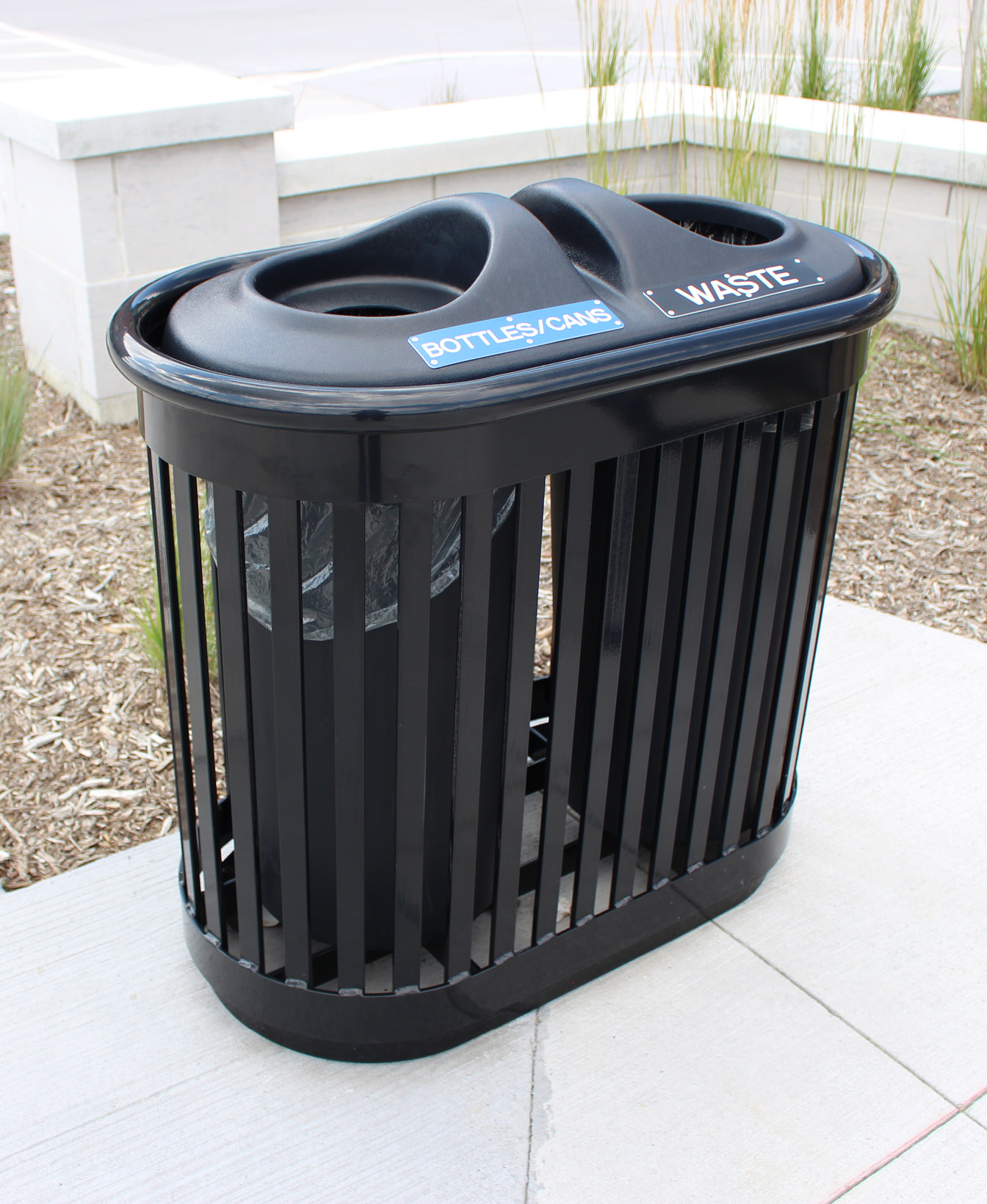 Le Contenant pour recyclage 250 est une poubelle extérieure noire avec des ouvertures séparées pour les "bouteilles/canettes" et les "déchets", placée sur un trottoir en béton près d'un aménagement paysager.