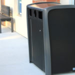 Une poubelle extérieure noire pour les déchets et le recyclage est placée sur un trottoir, près d'un banc et du mur d'un bâtiment.