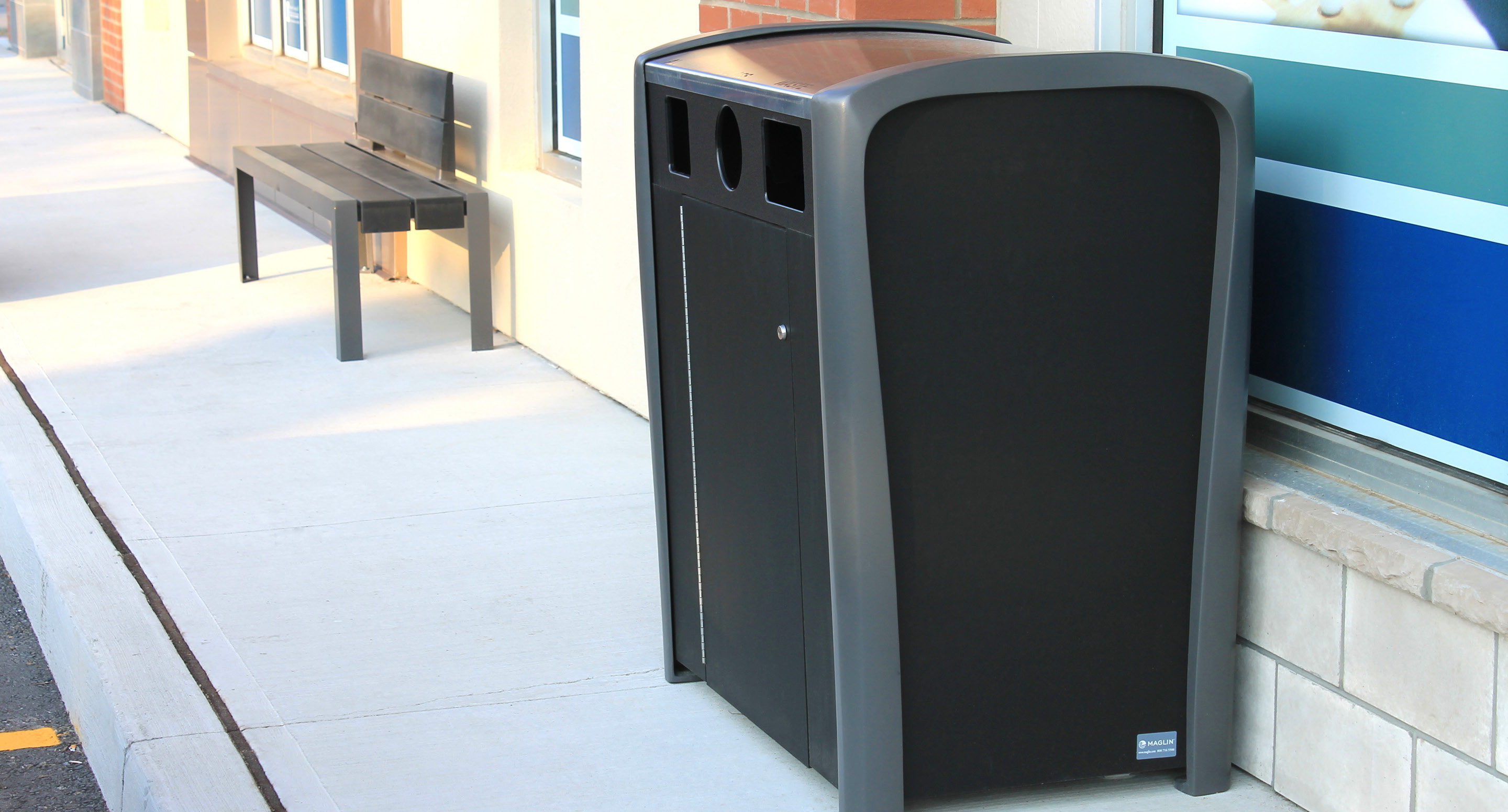 Une poubelle extérieure noire pour les déchets et le recyclage est placée sur un trottoir, près d'un banc et du mur d'un bâtiment.