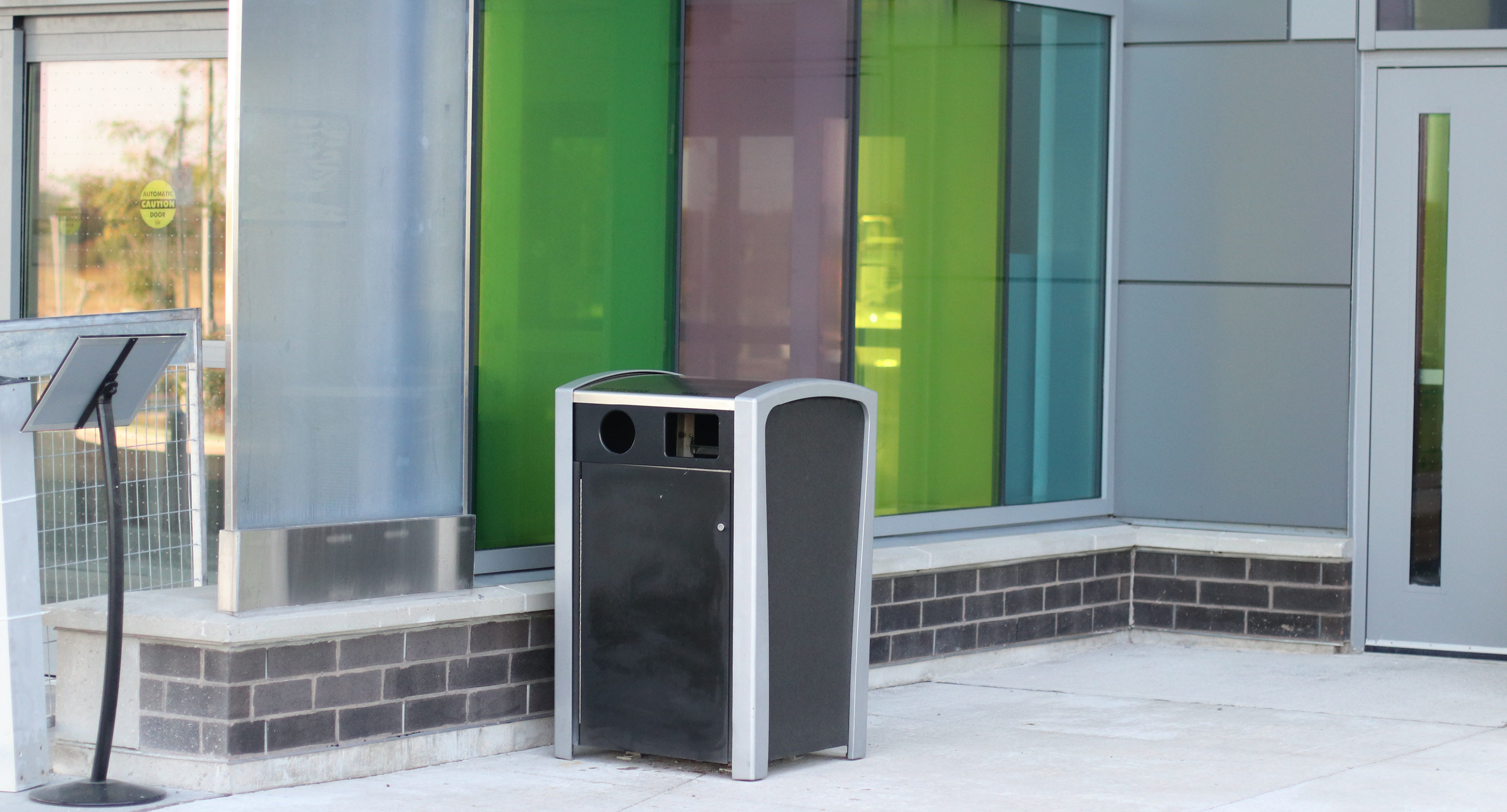 maglin_mrr-1400-recycle-receptacles-2-0613 Une poubelle extérieure noire et argentée est placée sur un trottoir devant un bâtiment aux panneaux de verre colorés.
