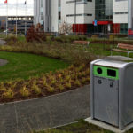 Une poubelle de recyclage et de déchets est située sur un chemin de gravier dans un espace extérieur aménagé avec des bancs, des plantes et un bâtiment moderne en arrière-plan.
