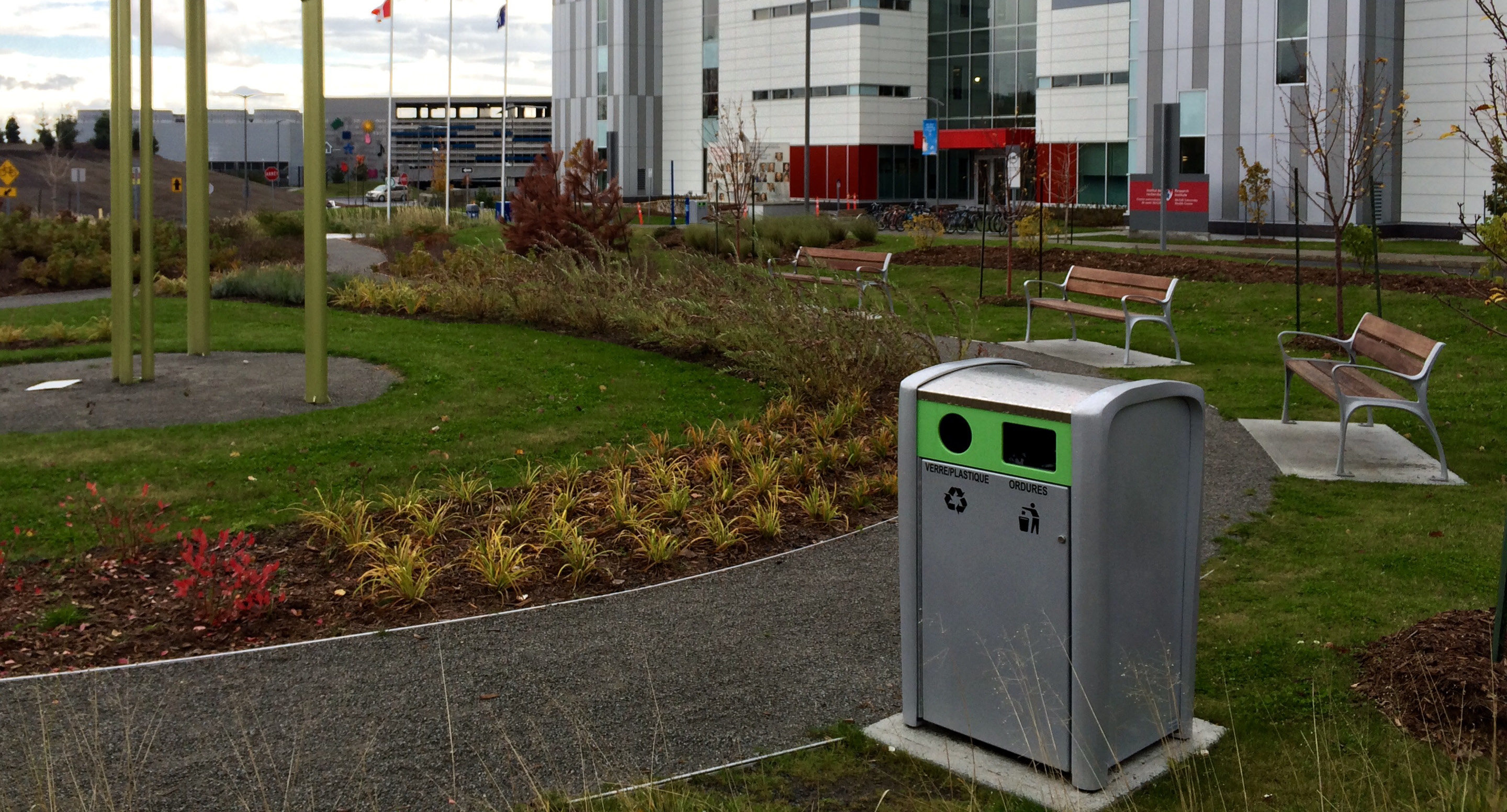 maglin_mrr-1400-recycle-receptacles-3 Une poubelle de recyclage et de déchets est située sur un chemin de gravier dans un espace extérieur aménagé avec des bancs, des plantes et un bâtiment moderne en arrière-plan.