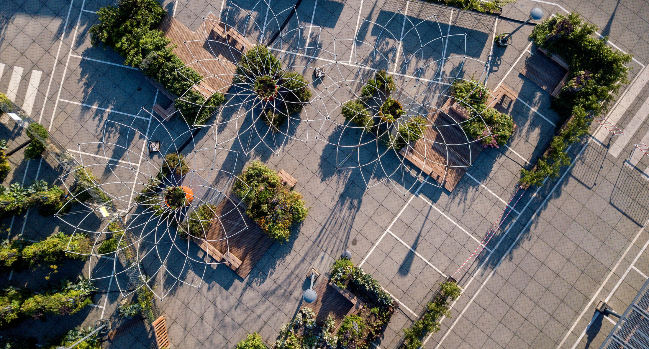 Aerial view of a parking lot converted into an urban garden with geometric wire structures, benches, and greenery arranged in patterned sections.