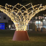 A large, cone-shaped outdoor light sculpture is illuminated at night on a grassy area, with buildings and string lights in the background.