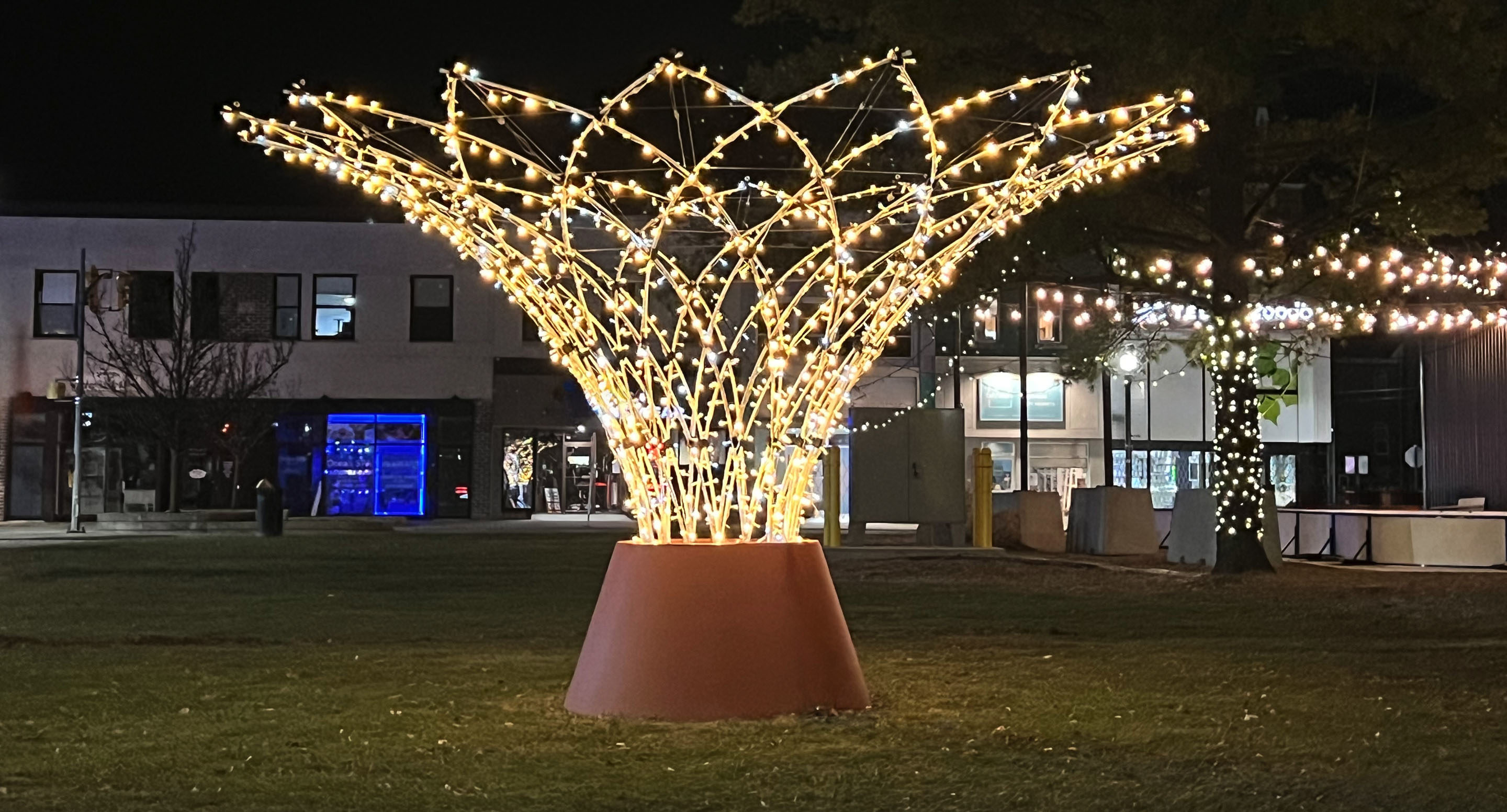 A large, cone-shaped outdoor light sculpture is illuminated at night on a grassy area, with buildings and string lights in the background.