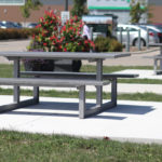 Table de pique-nique et banc gris posés sur une dalle en béton dans un espace extérieur herbeux, avec une autre table, des fleurs, des voitures garées et un bâtiment en arrière-plan.
