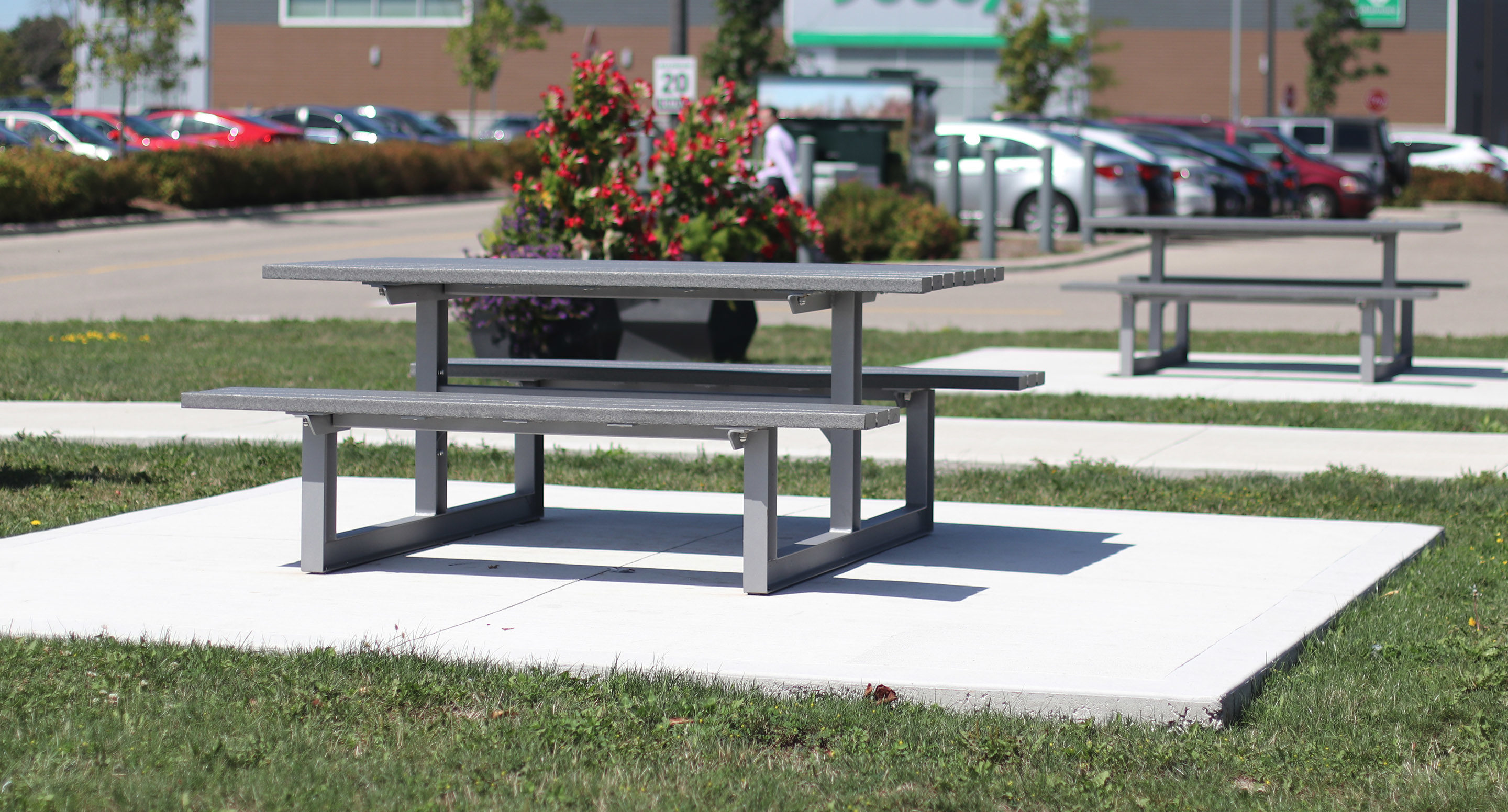 maglin_mtb-0210-cluster-seating-1 Table de pique-nique et banc gris posés sur une dalle en béton dans un espace extérieur herbeux, avec une autre table, des fleurs, des voitures garées et un bâtiment en arrière-plan.