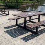 Deux tables de pique-nique en bois avec leurs bancs sont installées sur un patio en béton au bord d'un lac ; l'une d'entre elles est équipée d'un échiquier. Une clôture métallique sépare la terrasse de l'eau.