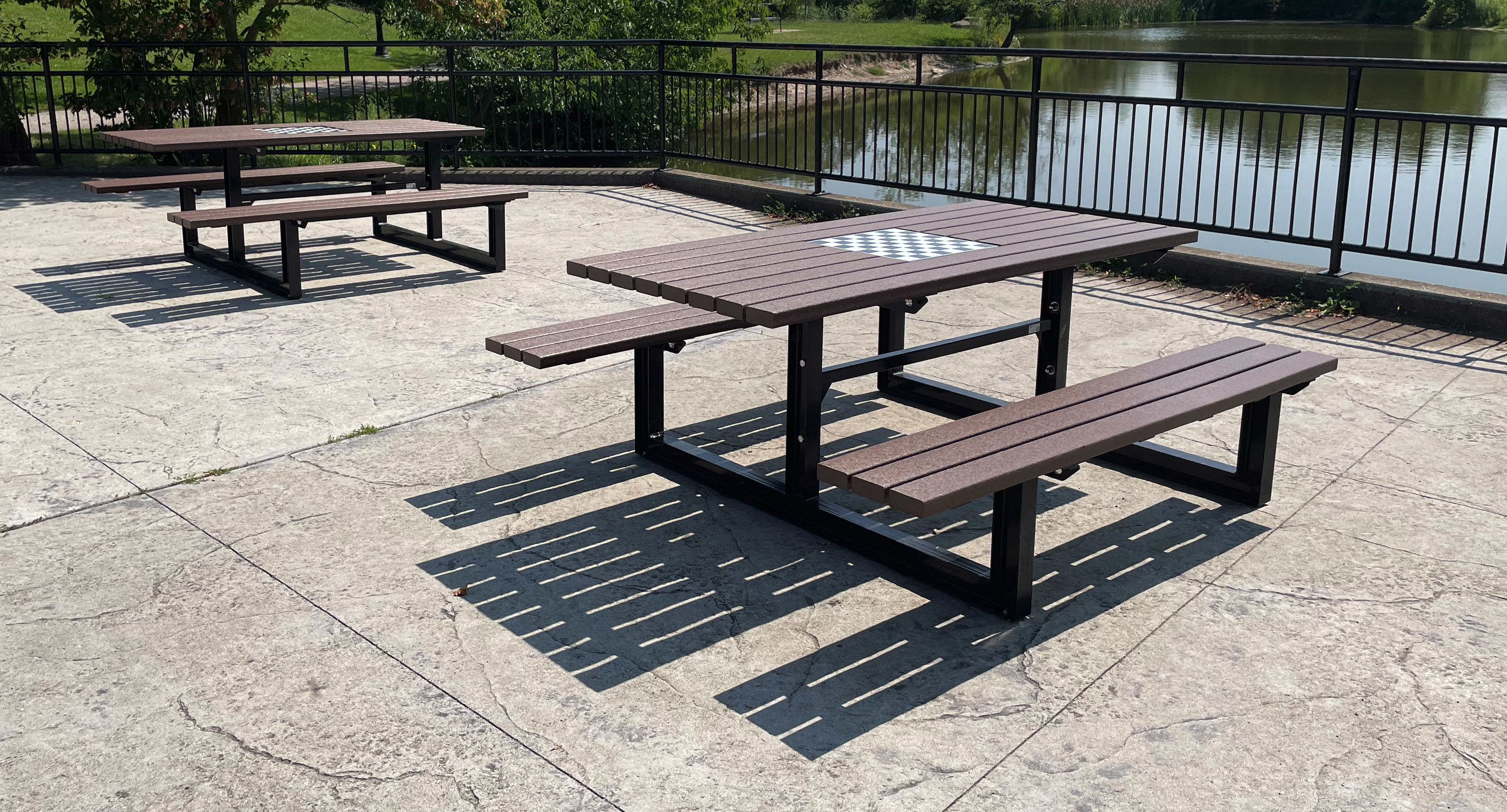 Deux tables de pique-nique en bois avec leurs bancs sont installées sur un patio en béton au bord d'un lac ; l'une d'entre elles est équipée d'un échiquier. Une clôture métallique sépare la terrasse de l'eau.