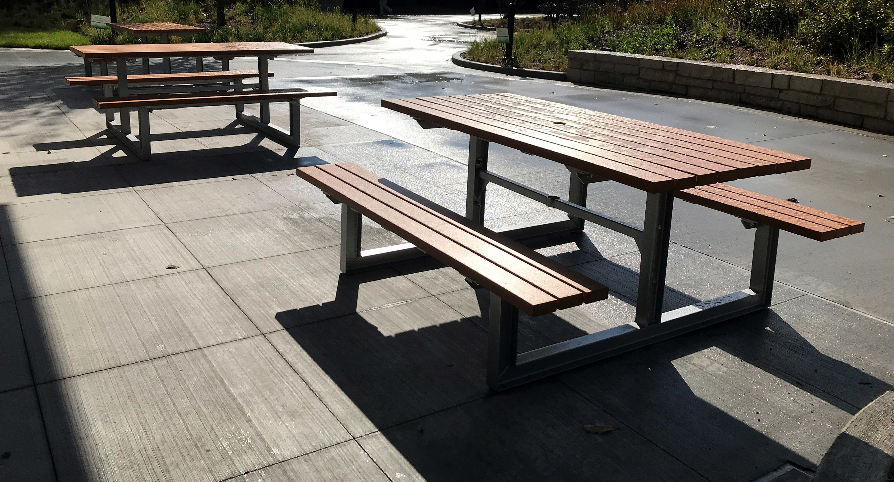 Deux tables de pique-nique vides en bois avec des cadres en métal sont placées sur un espace extérieur pavé ensoleillé, avec de la verdure et un mur en pierre à l'arrière-plan.