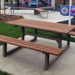 Une table de pique-nique en bois avec des bancs est posée sur une dalle en béton dans un parc près d'une aire de jeux avec des équipements colorés.