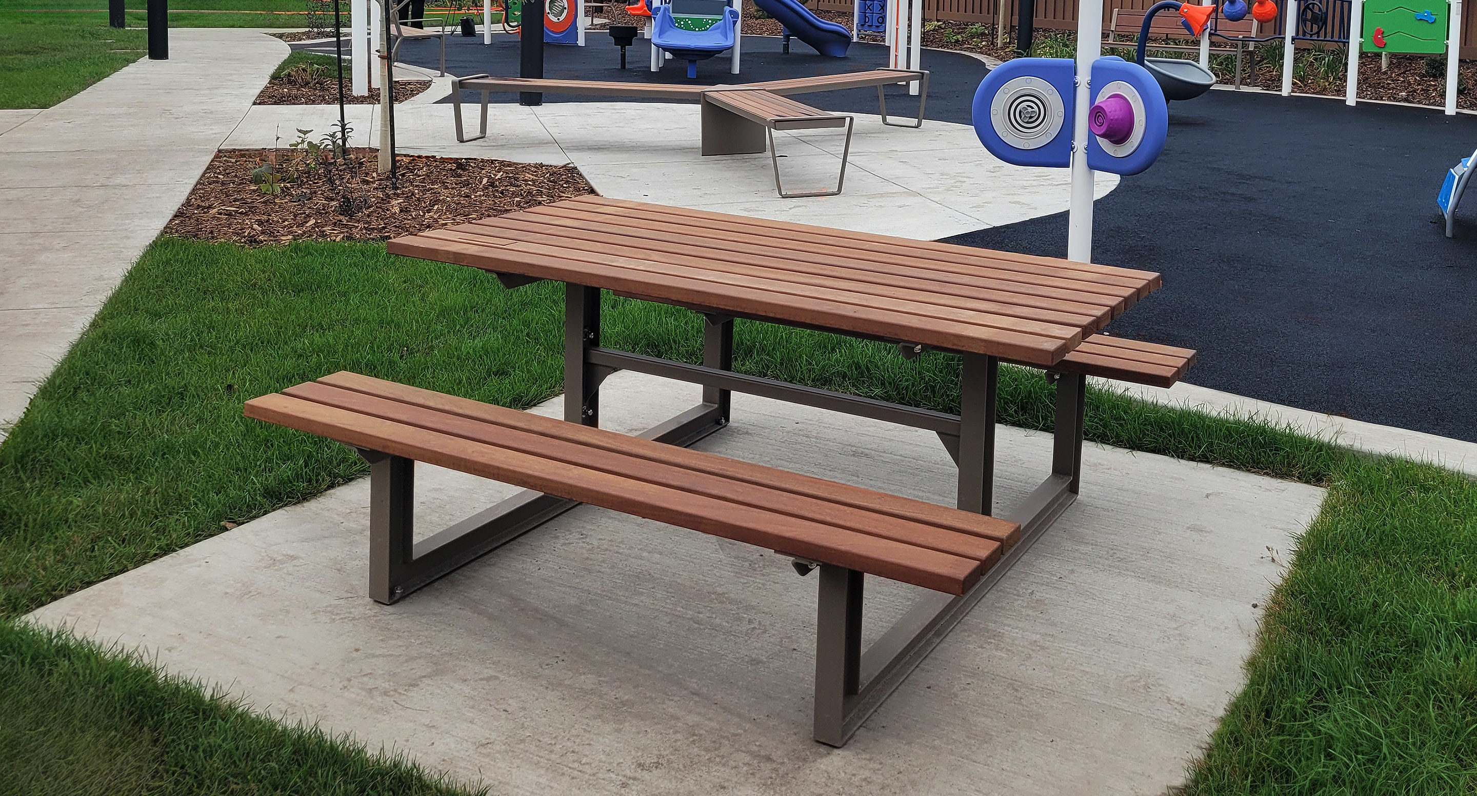 Une table de pique-nique en bois avec des bancs est posée sur une dalle en béton dans un parc près d'une aire de jeux avec des équipements colorés.