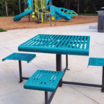 A teal metal picnic table with attached seats is on a concrete patio near a trash can, with a playground and another picnic table in the background.