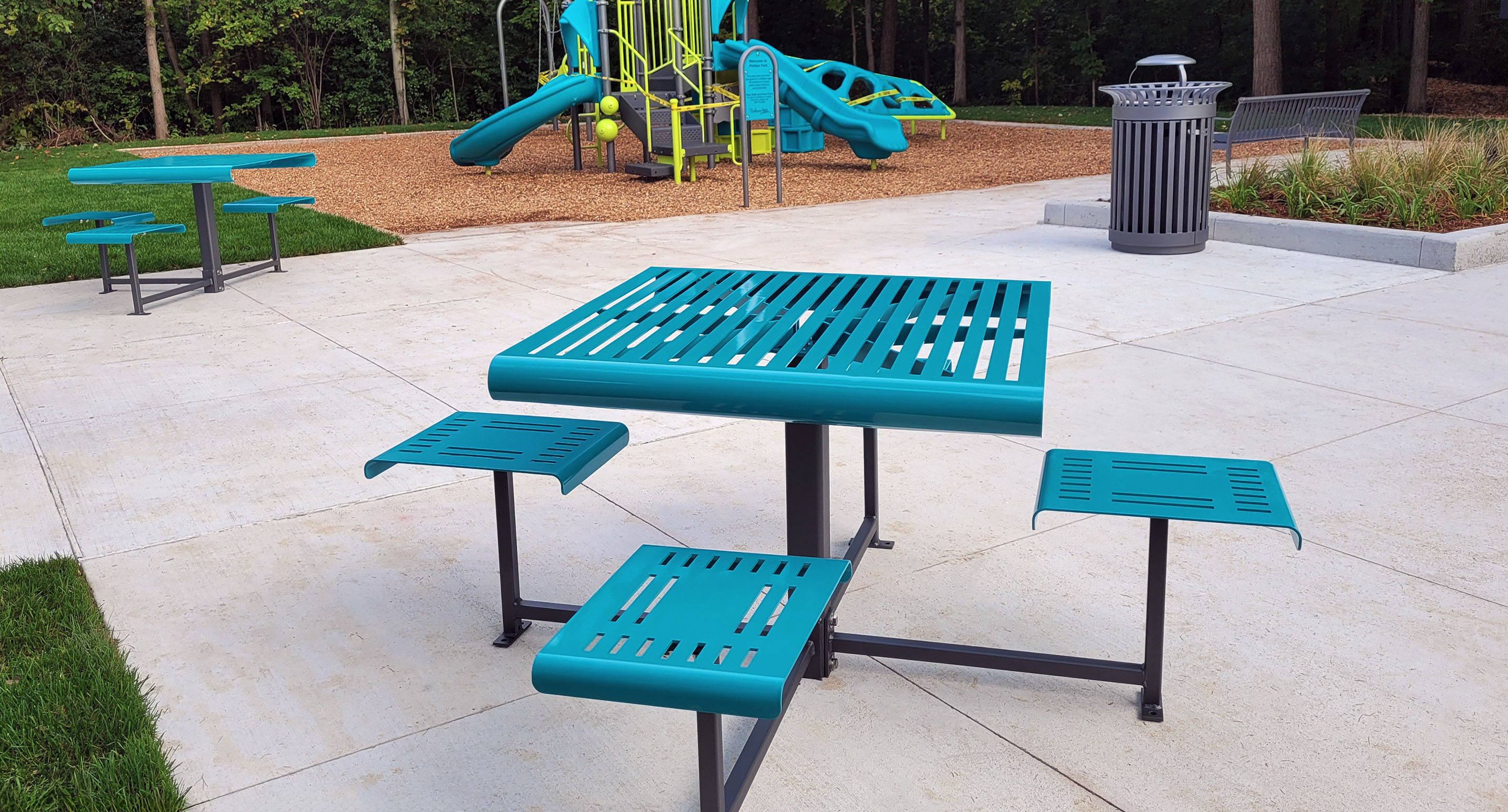 maglin_mtb-0400-cluster-seating-1 A teal metal picnic table with attached seats is on a concrete patio near a trash can, with a playground and another picnic table in the background.