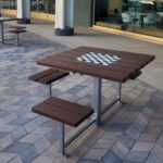 A brown outdoor table with built-in benches and a chessboard pattern on top sits on a paved patio near a building with large windows.