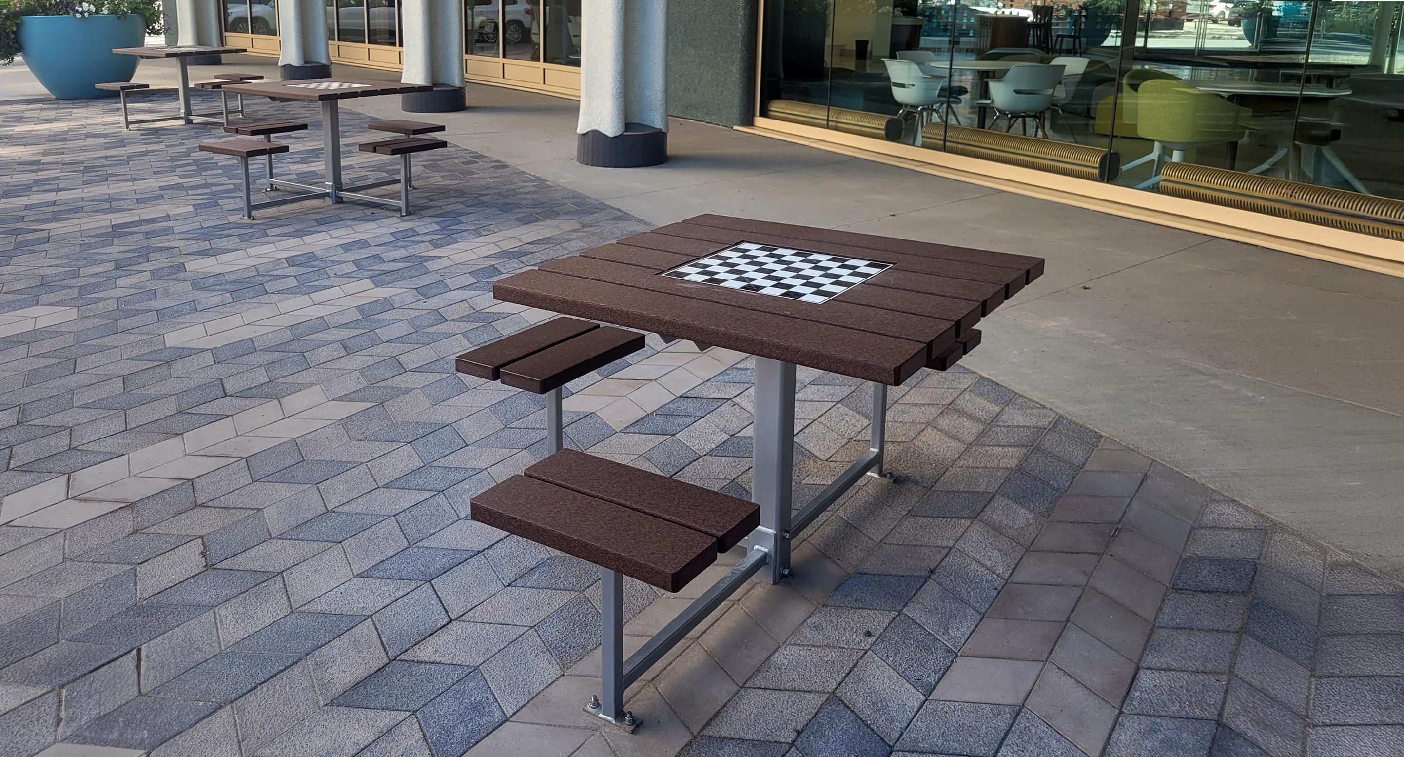maglin_mtb-0400-cluster-seating-2 A brown outdoor table with built-in benches and a chessboard pattern on top sits on a paved patio near a building with large windows.