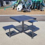 A metal outdoor table with two attached benches is placed on a concrete surface near a playground with climbing structures and slides.