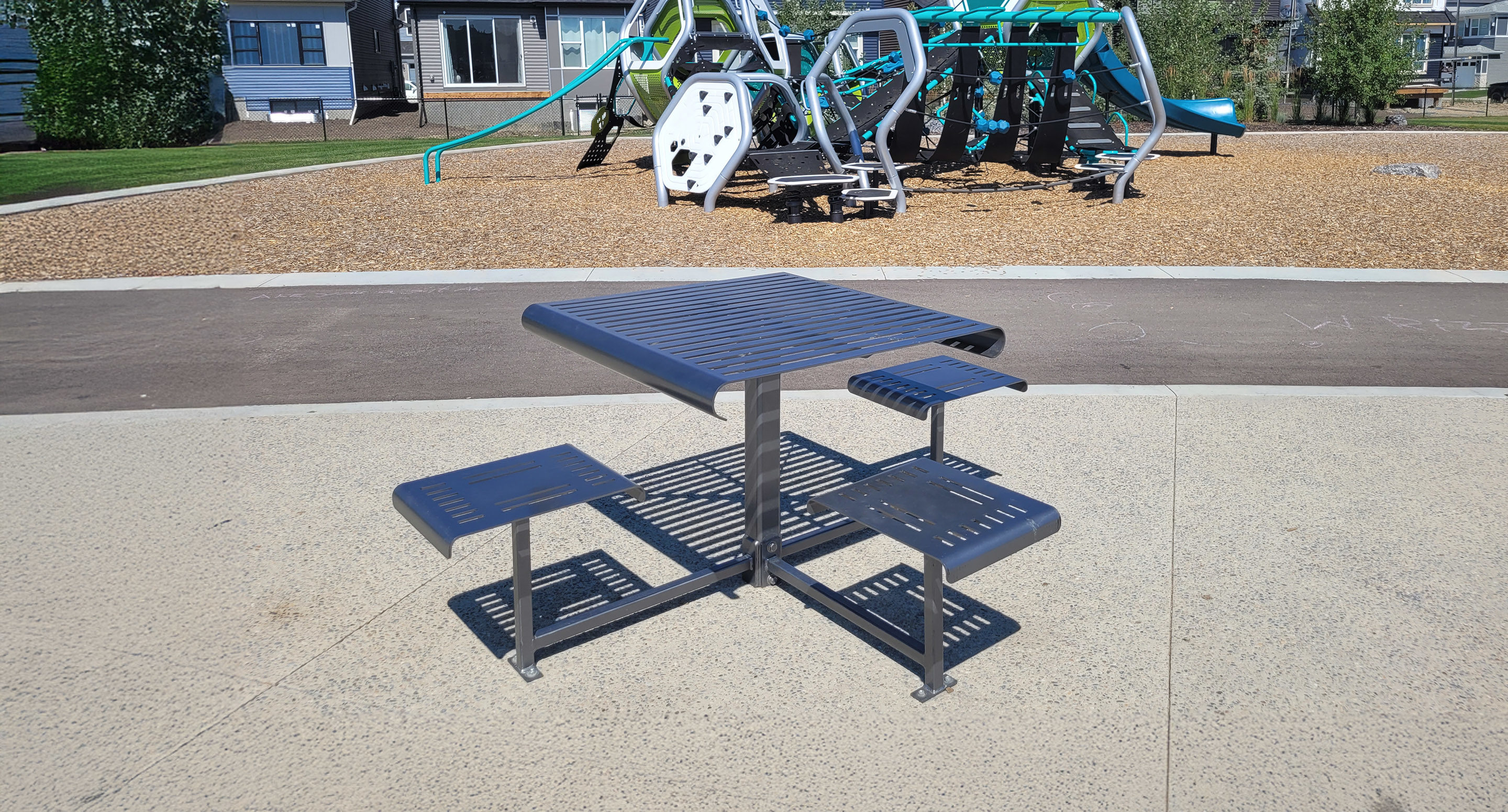maglin_mtb-0400-cluster-seating-3 A metal outdoor table with two attached benches is placed on a concrete surface near a playground with climbing structures and slides.