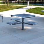 A modern metal picnic table with attached seats, a bench, and a ping pong table are situated on a concrete patio in a park.
