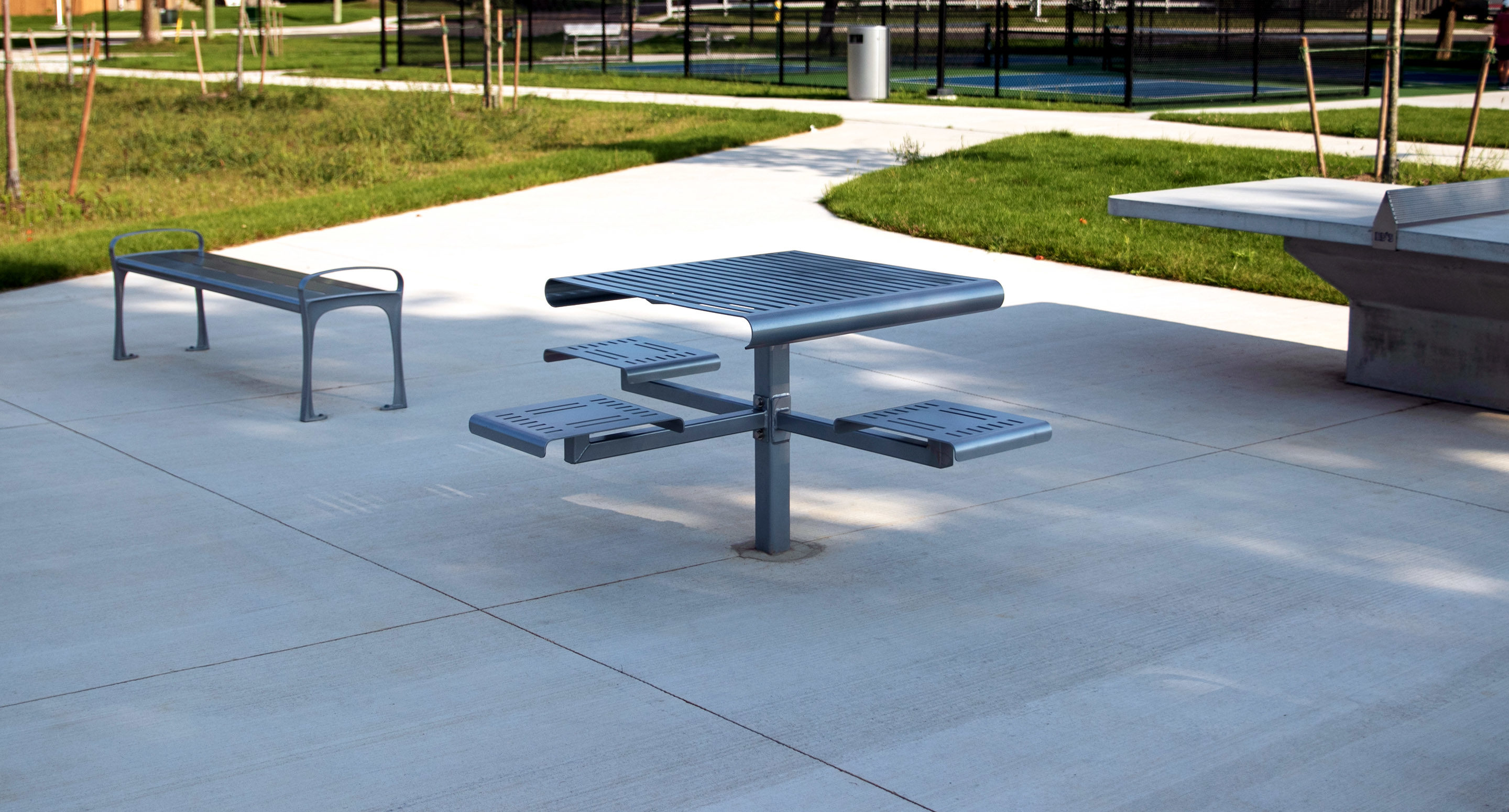 maglin_mtb-0400-cluster-seating-4 A modern metal picnic table with attached seats, a bench, and a ping pong table are situated on a concrete patio in a park.