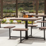 Outdoor picnic tables and benches with metal frames and wooden seats are arranged on a concrete patio, with landscaping and parked vehicles in the background.