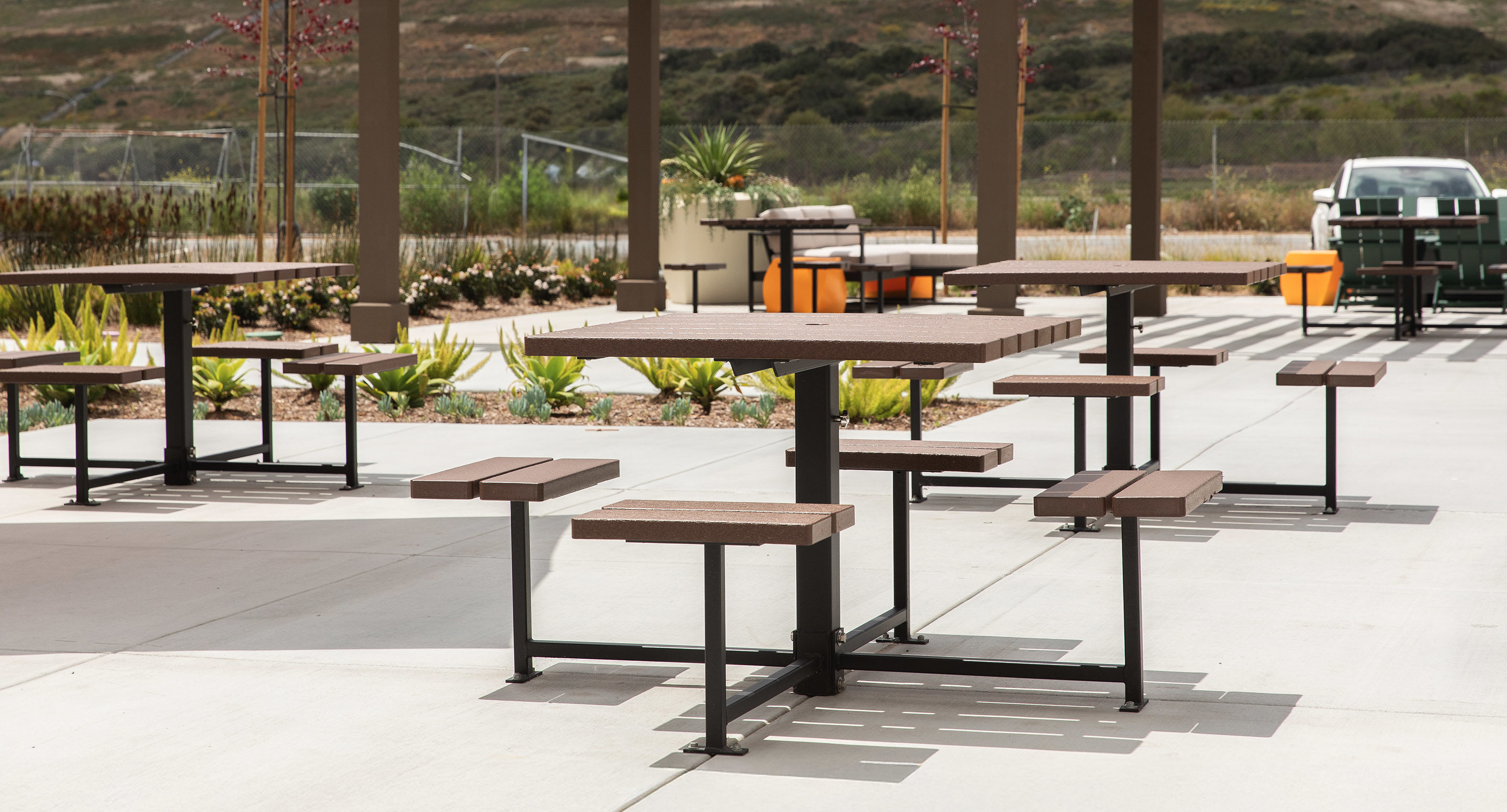maglin_mtb-0400-cluster-seating-5 Outdoor picnic tables and benches with metal frames and wooden seats are arranged on a concrete patio, with landscaping and parked vehicles in the background.
