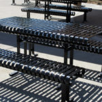 Tables de pique-nique et bancs en métal noir sur une surface en béton, projetant des ombres rayées en plein soleil.