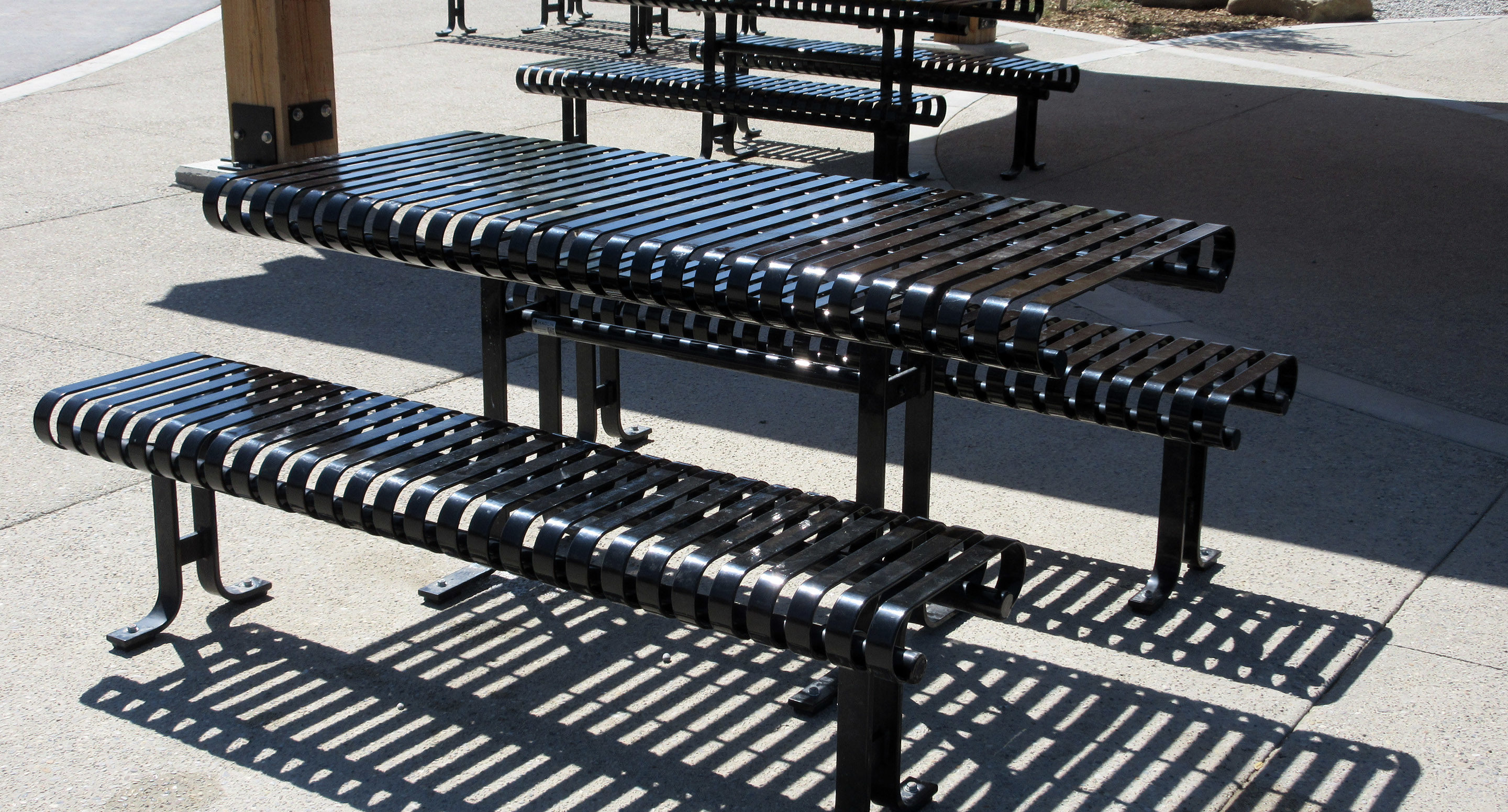 Tables de pique-nique et bancs en métal noir sur une surface en béton, projetant des ombres rayées en plein soleil.