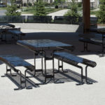 Espace extérieur couvert avec plusieurs tables de pique-nique et bancs en métal noir sur une surface en béton, entouré d'arbres, d'herbe et de quelques maisons en arrière-plan.
