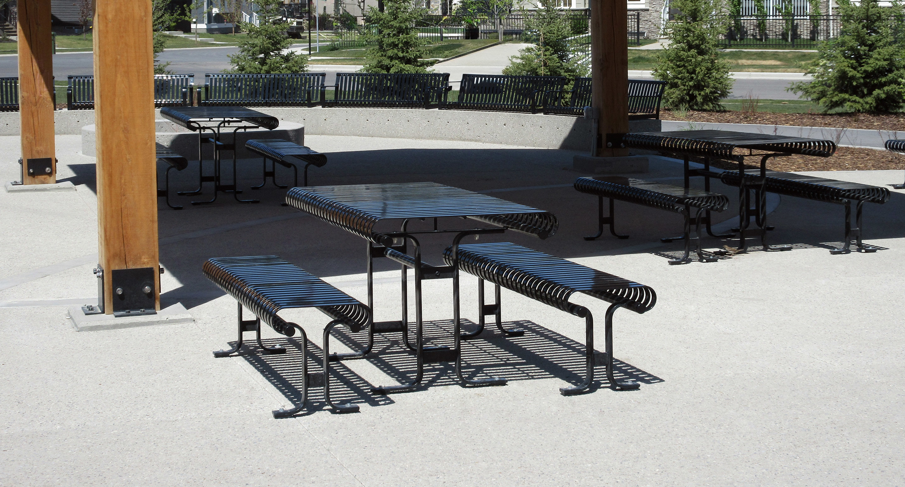 maglin_mtb-0510-cluster-seating-3 Espace extérieur couvert avec plusieurs tables de pique-nique et bancs en métal noir sur une surface en béton, entouré d'arbres, d'herbe et de quelques maisons en arrière-plan.
