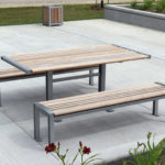 A modern outdoor picnic table with two matching benches, made of light wood and metal, is set on a concrete patio next to landscaping and a walkway.