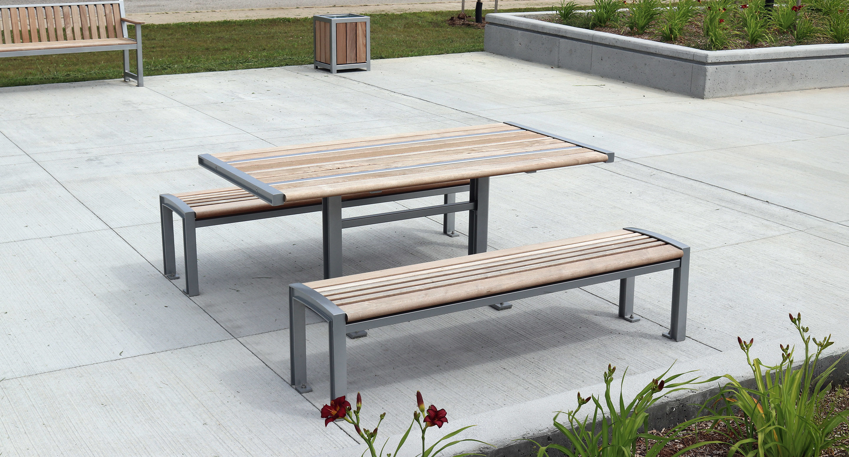 maglin_mtb-0720-cluster-seating-1 A modern outdoor picnic table with two matching benches, made of light wood and metal, is set on a concrete patio next to landscaping and a walkway.