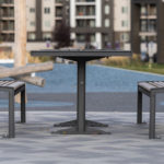 A metal outdoor table with two matching benches is set on a paved area in a park, with apartment buildings in the background.