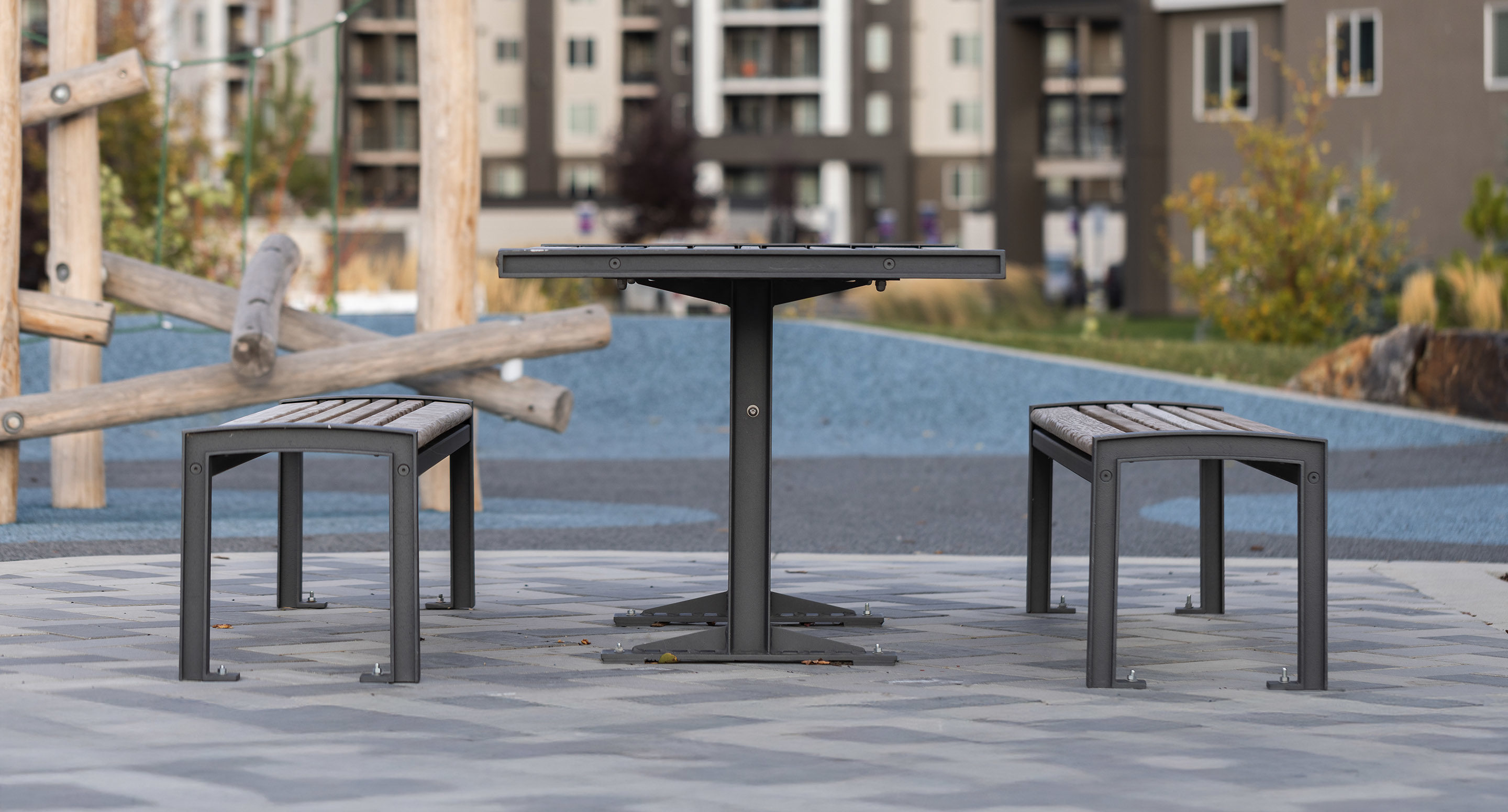 maglin_mtb-0720-cluster-seating-2 A metal outdoor table with two matching benches is set on a paved area in a park, with apartment buildings in the background.