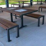 Outdoor picnic tables and benches with metal frames and wooden seats, one table featuring a built-in chessboard, set on a concrete surface under a covered area.