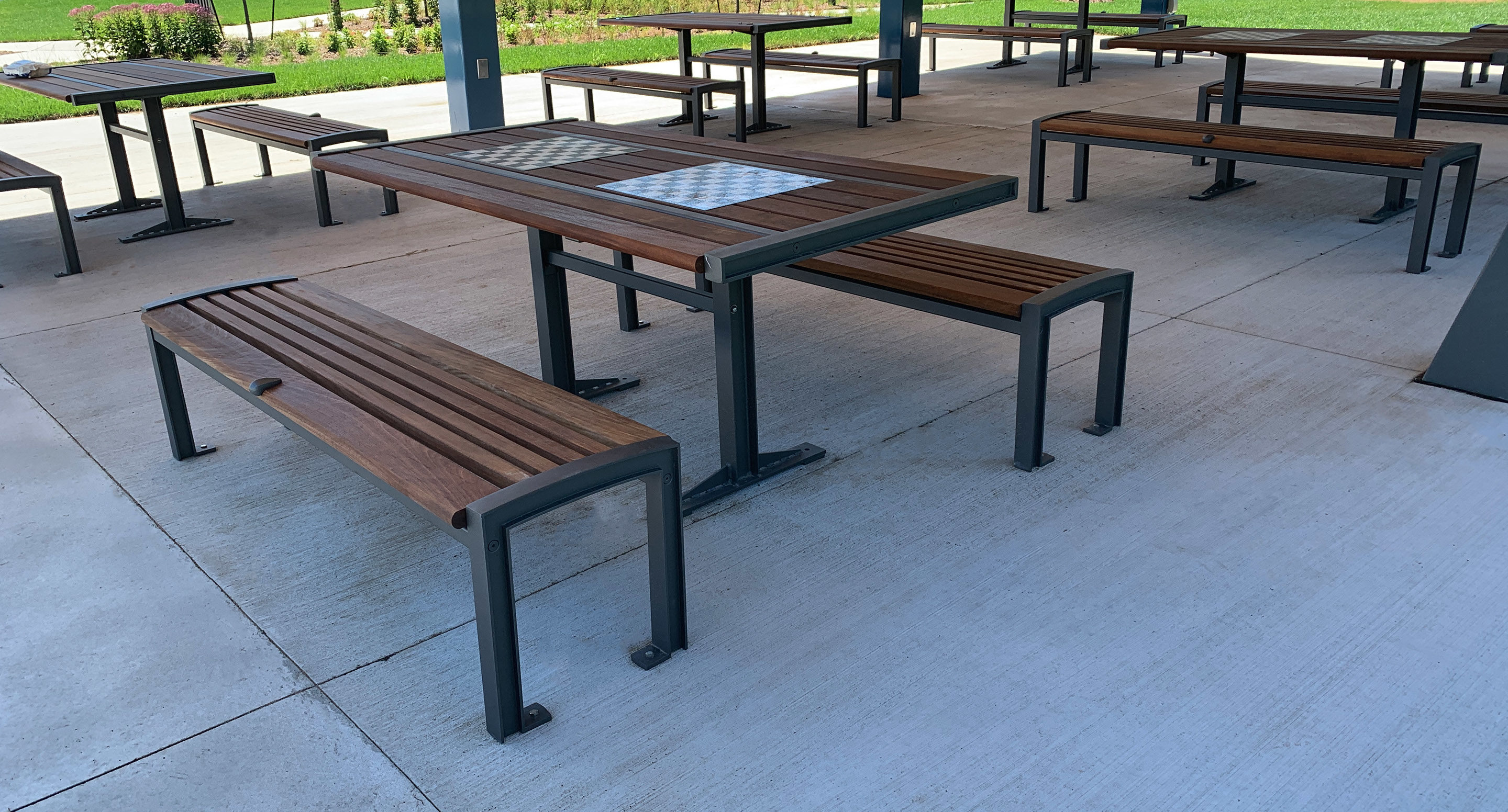 maglin_mtb-0720-cluster-seating-3 Outdoor picnic tables and benches with metal frames and wooden seats, one table featuring a built-in chessboard, set on a concrete surface under a covered area.
