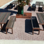 Four wooden picnic tables with matching benches are arranged on a brick patio, with a tree in the center and dry, brown dirt areas surrounding the patio.