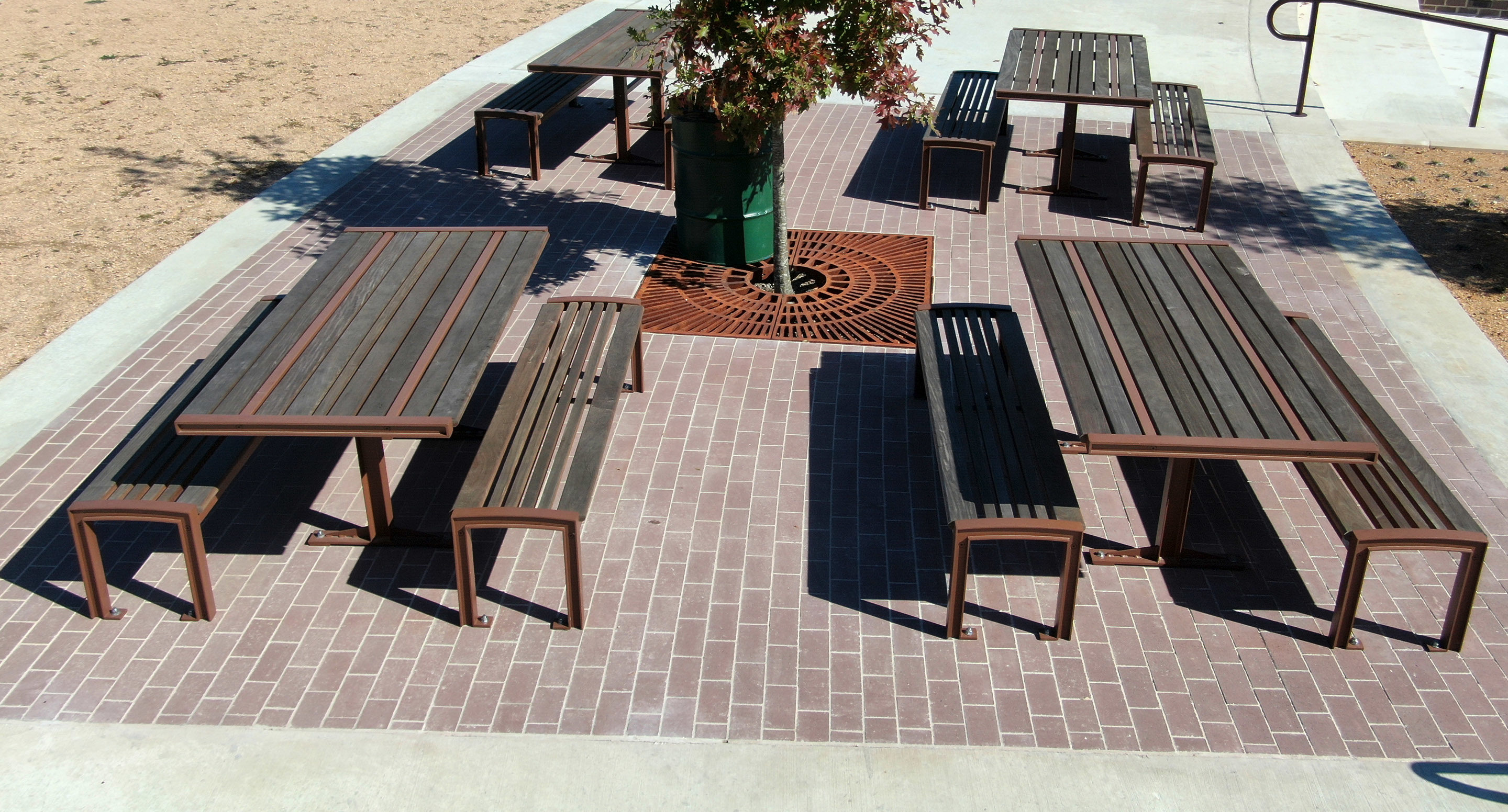 maglin_mtb-0720-cluster-seating-4 Four wooden picnic tables with matching benches are arranged on a brick patio, with a tree in the center and dry, brown dirt areas surrounding the patio.