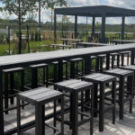 Outdoor patio area with rows of black tables and stools on a paved surface, surrounded by greenery and a fenced boundary under a partly cloudy sky.